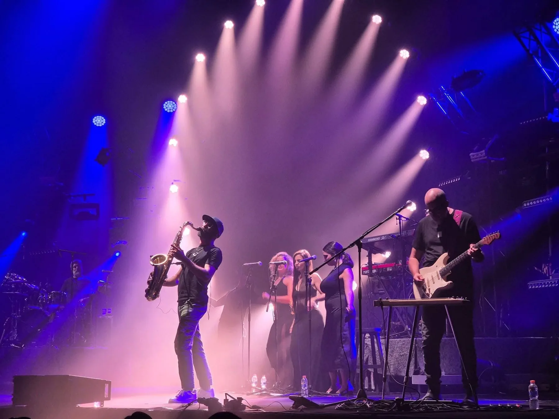 Echoes on stage: saxophone, three backing vocalists and guitar under white spotlights